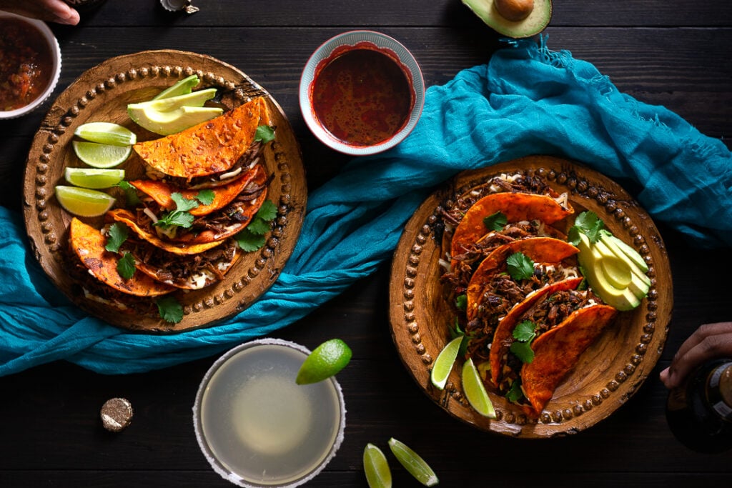 A wooden table setting with 2 wooden plates with 3 tacos each and avocado slices, sides of salsa & birria consomme. One margarita with lime wedge. Blue table cloth