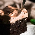 slice of chocolate cake topped with milk chocolate ganache on white plate with silver fork about to go through slice