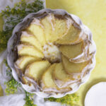 sliced lemon bundt cake with lemon sugar coating and lemon frosting glaze on a white cake stand with slices arranged in a circular domino pattern