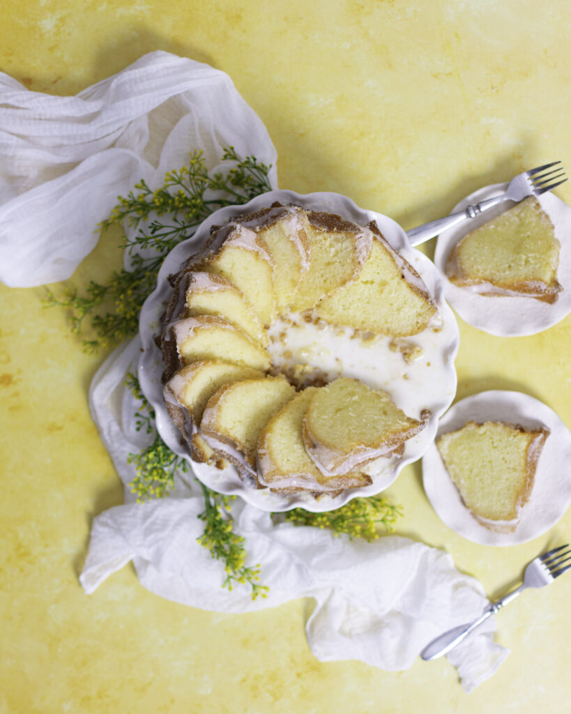 sliced lemon bundt cake with lemon sugar coating and lemon frosting glaze on a white cake stand with slices arranged in a circular domino pattern. 2 small tea plates to the right of cake stand with 1 slice of lemon bundt cake and a silver fork on each plate
