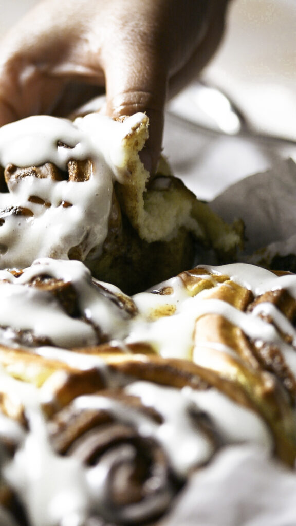 hand lifting brioche style cinnamon rolls with cream cheese frosting out of parchment paper-lines cast iron pan