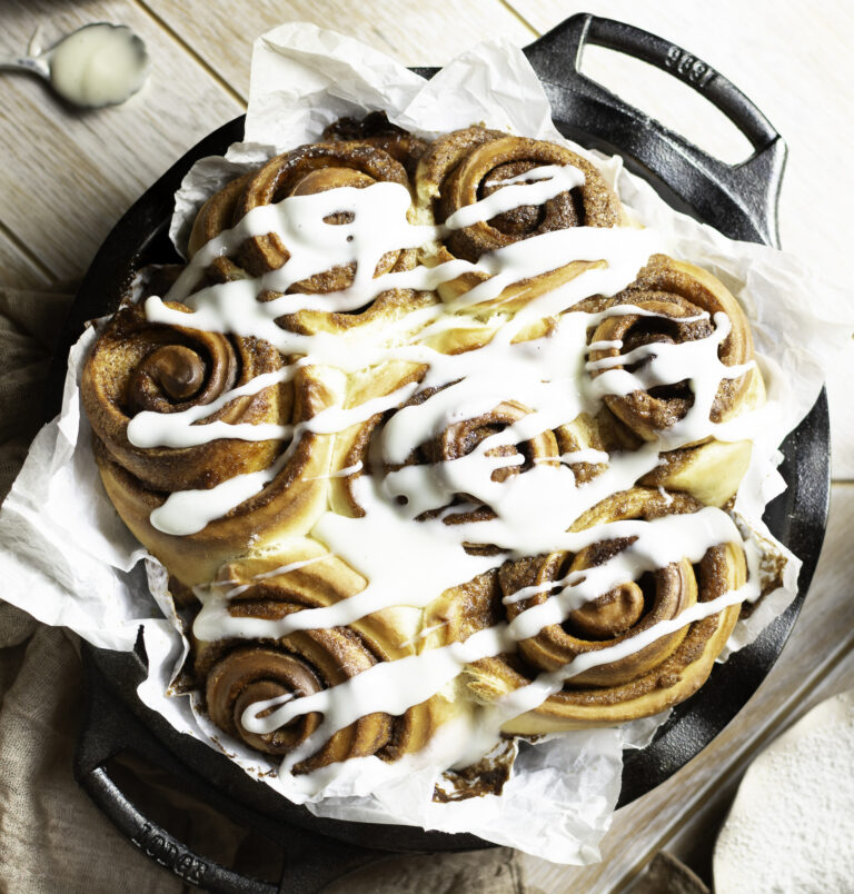 parchment-lined cast iron pan filled with cinnamon rolls and drizzled with cream cheese frosting