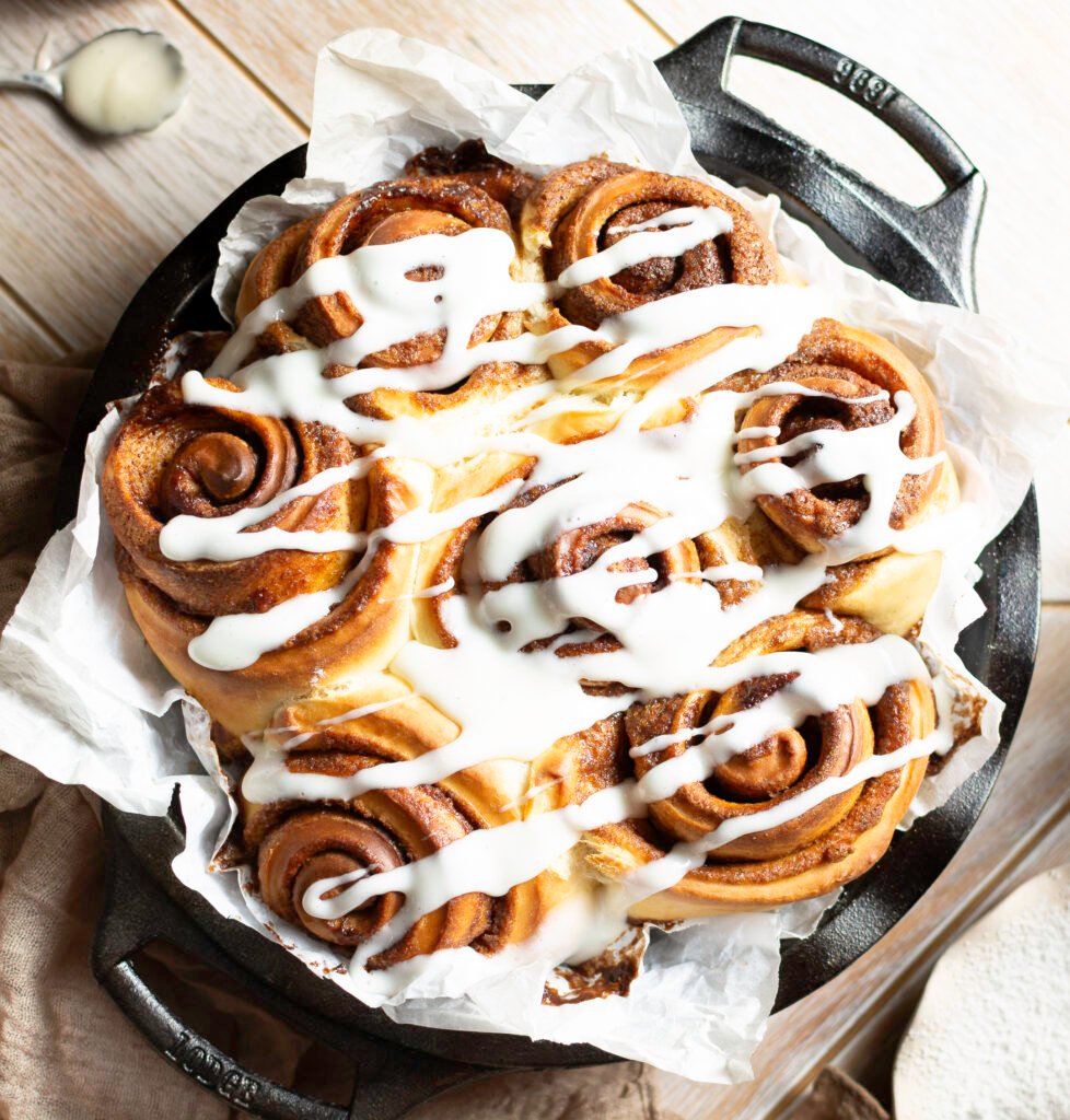parchment-lined cast iron pan filled with cinnamon rolls and drizzled with cream cheese frosting