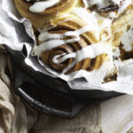 brioche-style cinnamon rolls in a parchment paper-lined Lodge cast iron pan drizzled with cream cheese frosting.