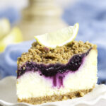 slice of lemon blueberry crumb cheesecake on plate with a lemon wedge on top of slice