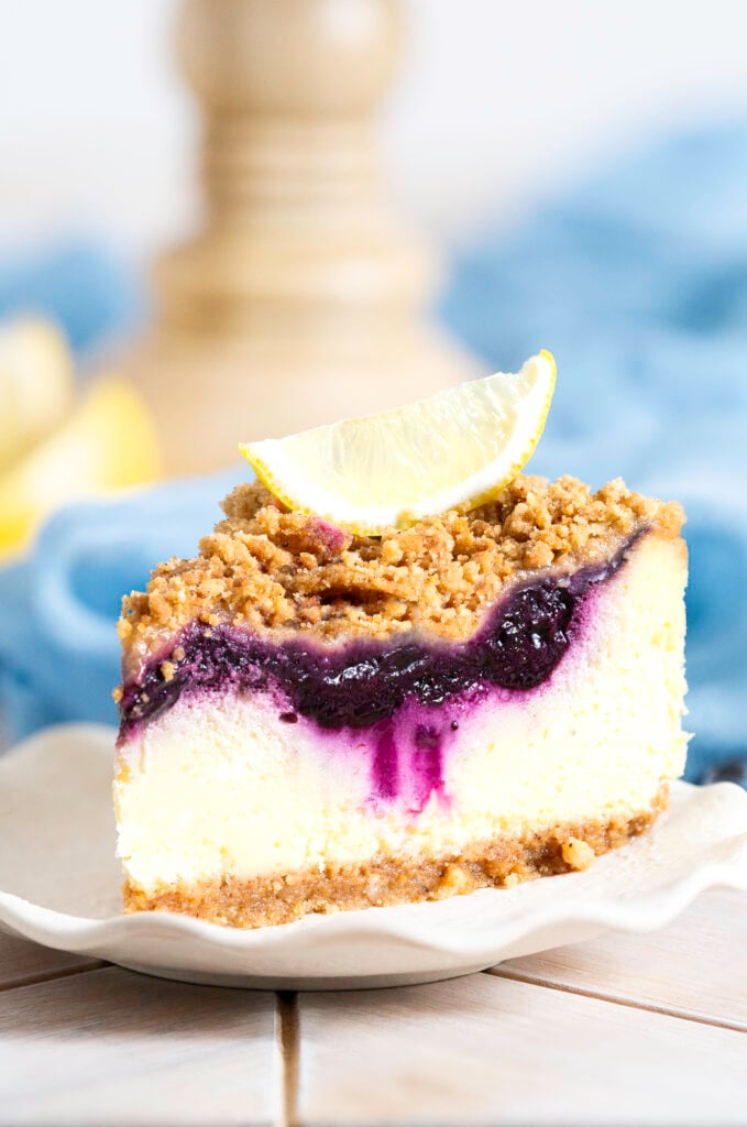 slice of lemon blueberry crumb cheesecake on plate with a lemon wedge on top of slice
