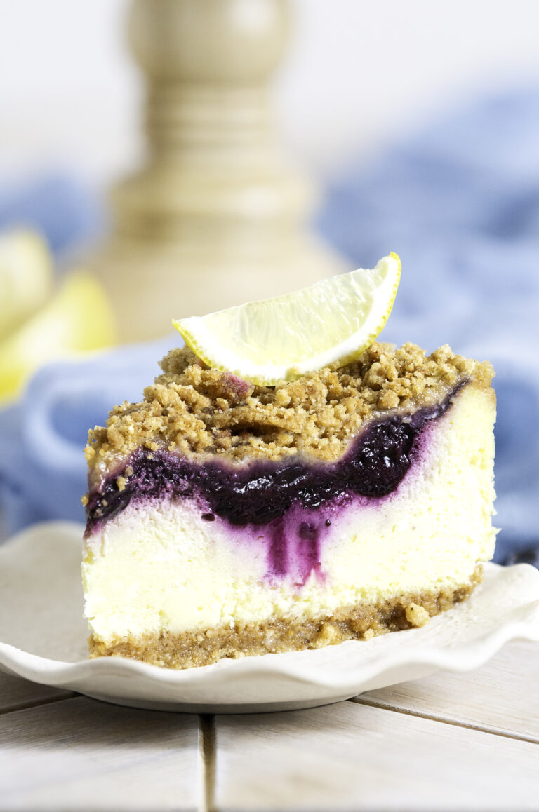 slice of lemon blueberry crumb cheesecake on plate with a lemon wedge on top of slice