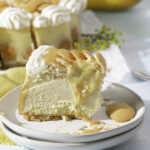 side profile of a slice of baked banana pudding cheesecake topped with sliced bananas, custard, Nilla wafers and whipped cream. whole banana pudding cheesecake in background.