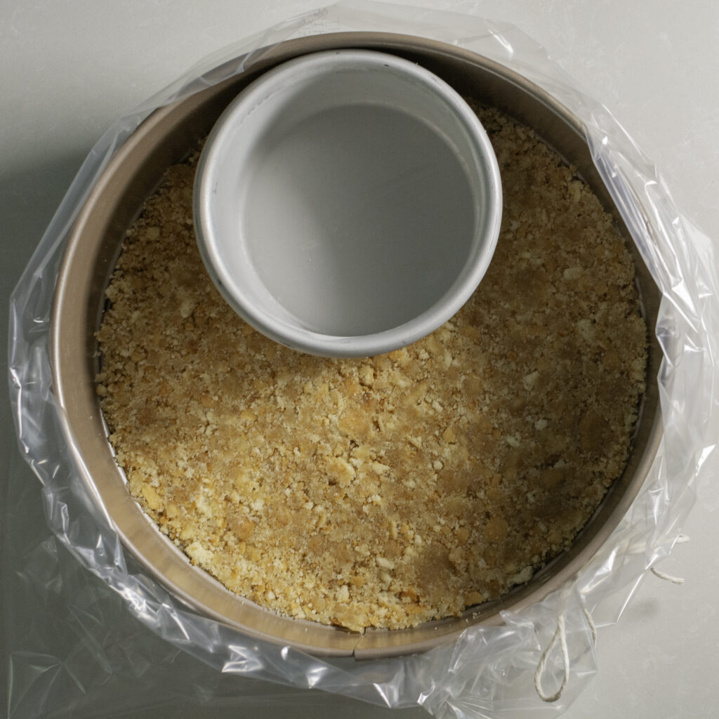 Nilla wafer cheesecake crust being pressed into the bottom a springform pan with smaller cake pan