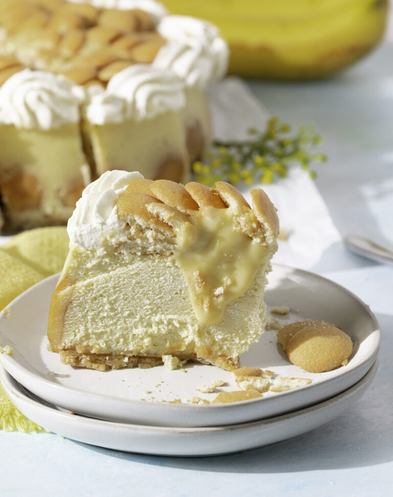 side profile of a slice of baked banana pudding cheesecake topped with sliced bananas, custard, Nilla wafers and whipped cream. whole banana pudding cheesecake in background.