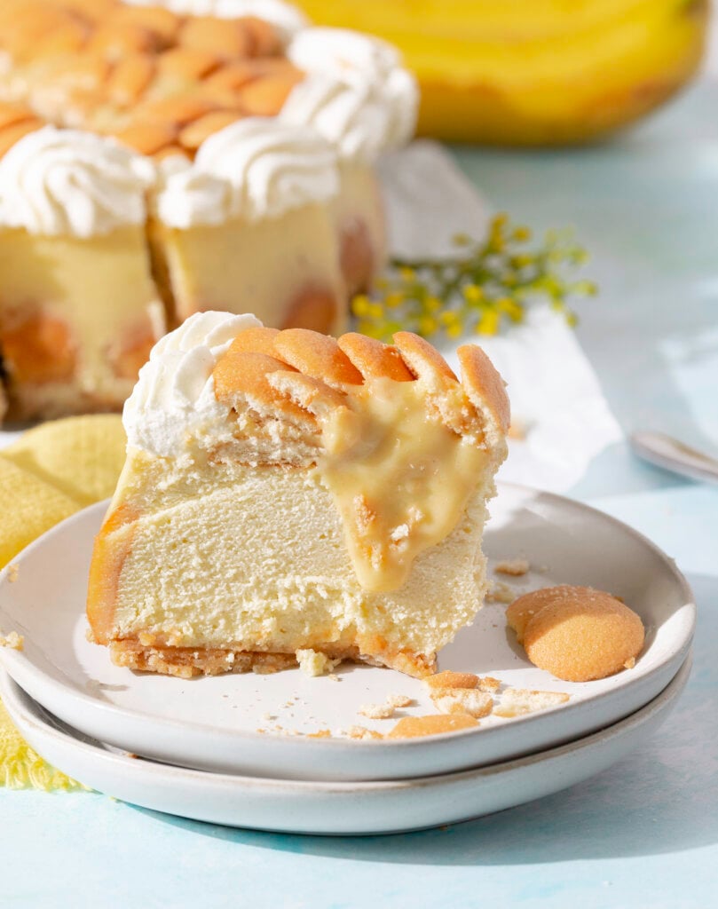 side profile of a slice of baked banana pudding cheesecake topped with sliced bananas, custard, Nilla wafers and whipped cream. whole banana pudding cheesecake in background.