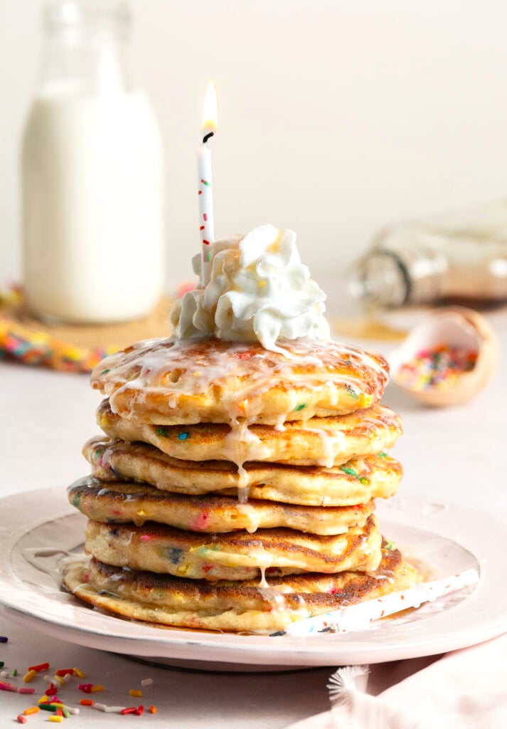 stack of 6 funfetti birthday buttermilk pancakes topped with whipped cream, confectioner's sugar glaze and lit birthday candle