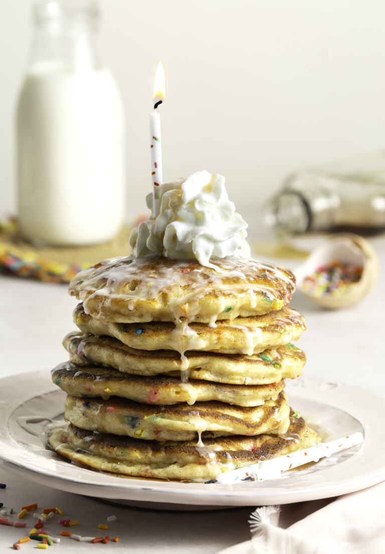 stack of 6 funfetti birthday buttermilk pancakes topped with whipped cream, confectioner's sugar glaze and lit birthday candle