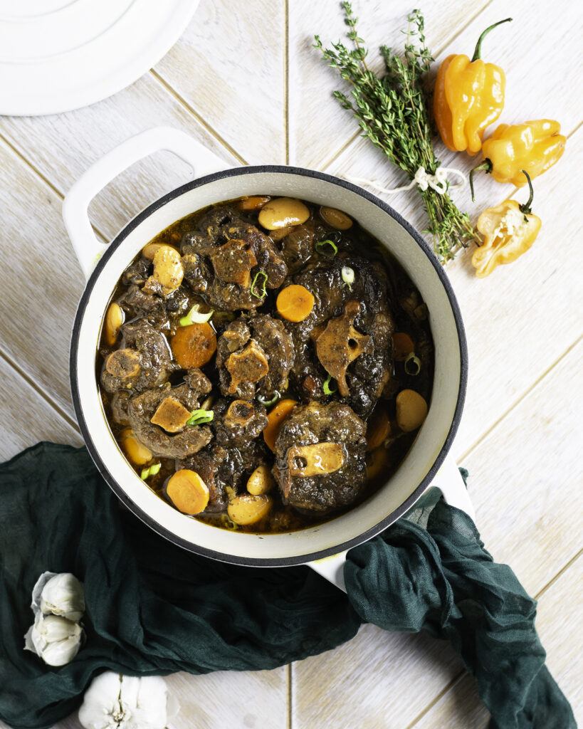 dutch oven filled with Jamaican oxtail with a bouquet of fresh thyme, scotch bonnet peppers and a bulb of garlic next to pot