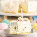 slice of no-bake white chocolate funfetti cheesecake on w white plate with a confetti candle on top and a bte missing from cheesecake