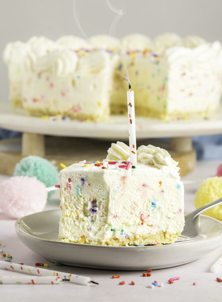 slice of no-bake white chocolate funfetti cheesecake on w white plate with a confetti candle on top and a bte missing from cheesecake