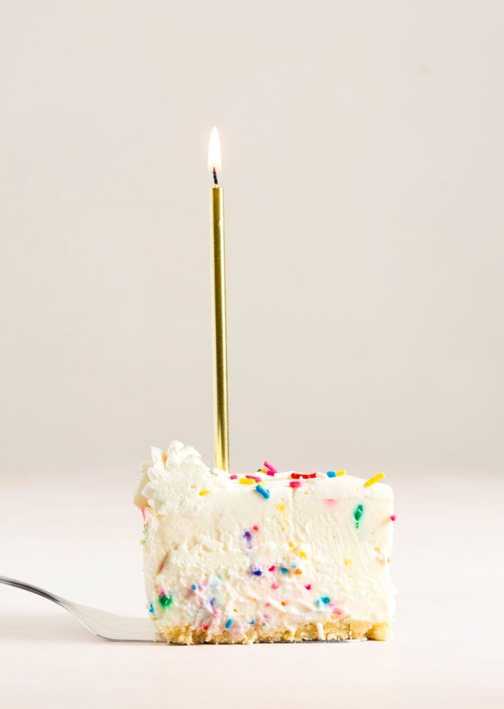 slice of no-bake white chocolate funfetti cheesecake on a cake server with a gold lit candle on top