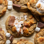 brown butter s'mores cookies on brown parchment paper with one cookie broken in half showing marshmallow stretch