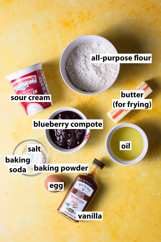 ingredients for homemade blueberry pancakes - all-purpose flour, sour cream, blueberry compote, oil, egg, vanilla extract, baking powder, baking soda, salt & butter for frying
