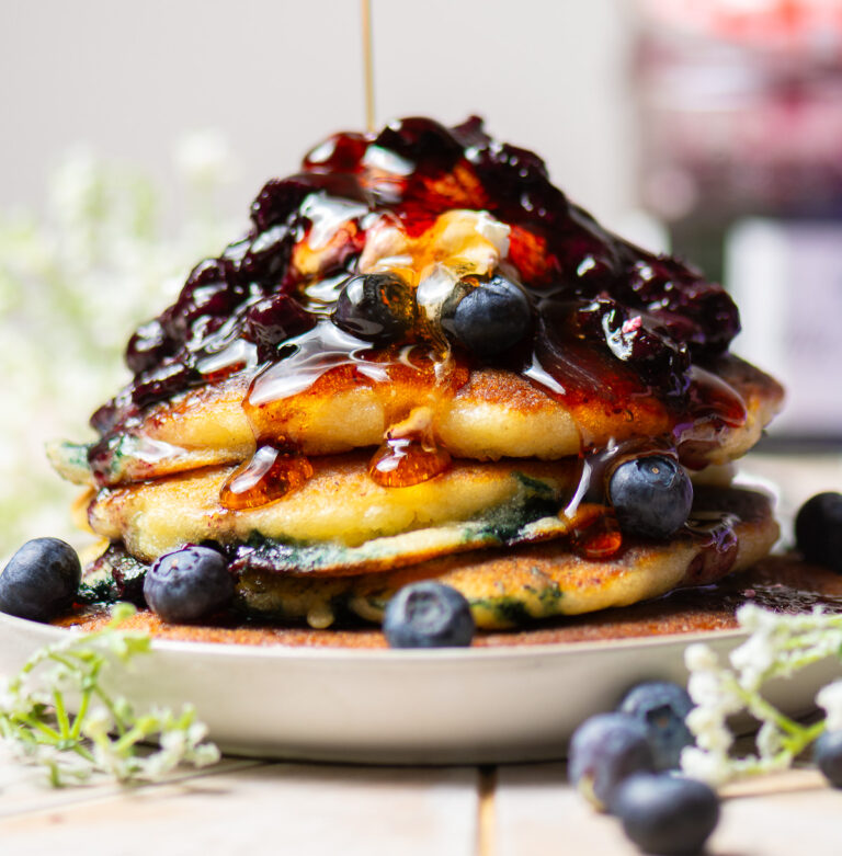 stack of homemade blueberry pancakes on a plate topped with fresh blueberry compote, cool whip & maple syrup