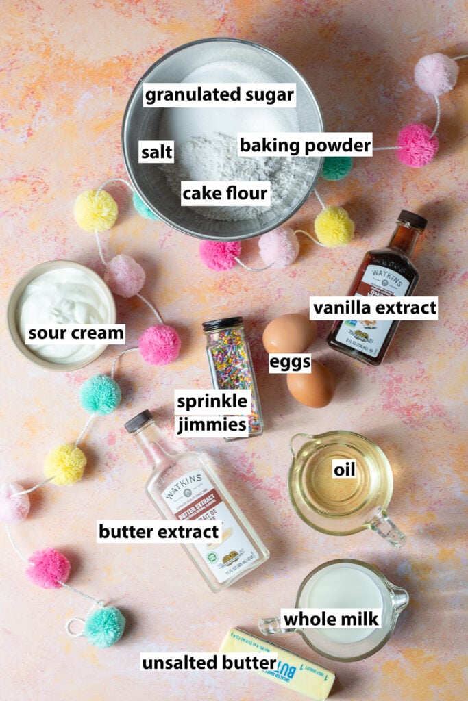 ingredients for a funfetti snack cake - cake flour, granulated sugar, baking powder, salt, vanilla extract, eggs, sprinkle jimmies, sour cream, oil, butter, milk, vanilla extract and butter extract