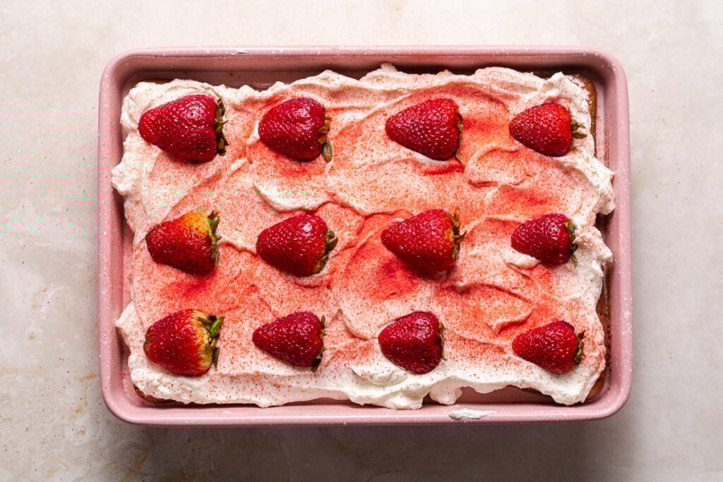 strawberry tres leches cake in a pink pan garnished with a dusting of freeze-dried strawberries and halved fresh strawberries