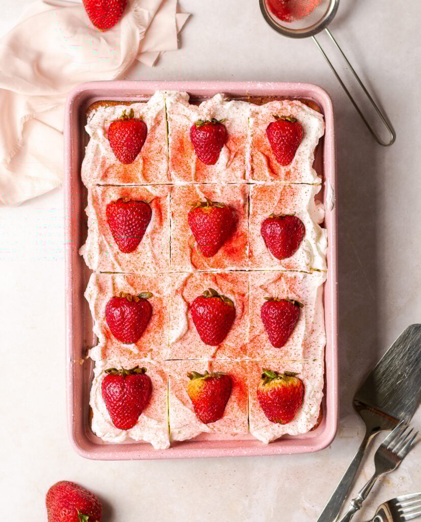 strawberry tres leches cake in a pink pan garnished with a dusting of freeze-dried strawberries and halved fresh strawberries