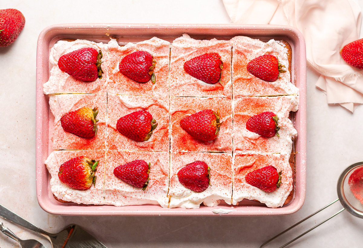 strawberry tres leches cake in a pink pan garnished with a dusting of freeze-dried strawberries and halved fresh strawberries