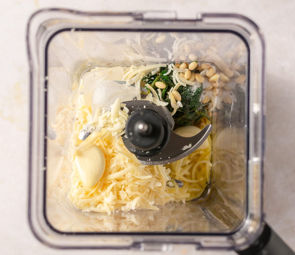 ingredients for classic basil pesto in a blender - basil, pine nuts, garlic, olive oil, parmesan cheese, pecorino romano cheese & ice cubes