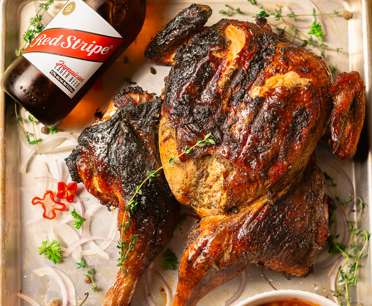 whole jerk chicken on a metal baking tray with a bottle of Red Stripe beer