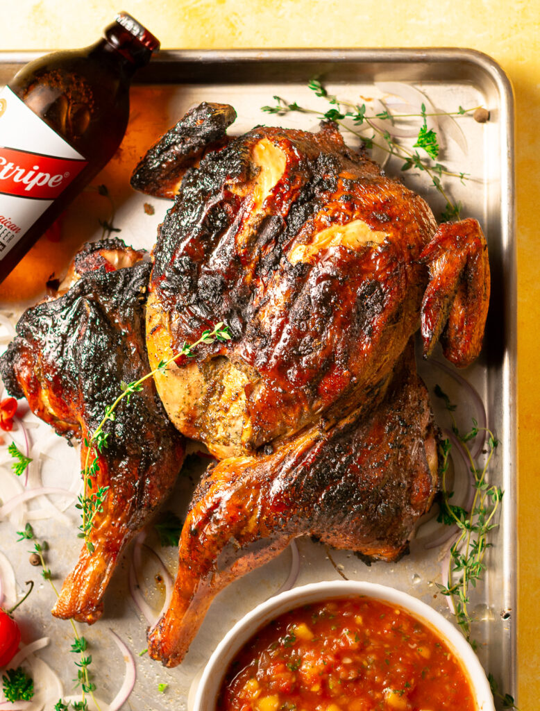 whole jerk chicken on a metal baking tray with a bottle of Red Stripe beer and bowl of mango habanero salsa