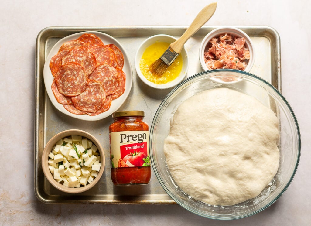 ingredients to assemble homemade pizza - homemade pizza dough, pizza sauce, mozzarella, prosciutto, garlic olive oil & chorizo