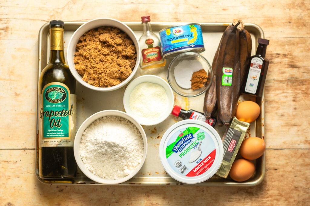 ingredients for hummingbird bundt cake - bananas, canned crushed pineapples, pecans, whole milk Greek yogurt, dark brown sugar, baking powder, baking soda, cinnamon, salt, eggs, butter, milk powder, dark rum, vegetable oil, vanilla extract and coconut extract
