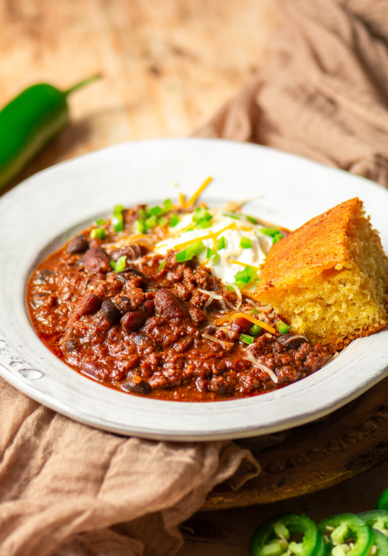 beef and chorizo chili in a bowl topped with sour cream, shredded cheese, diced jalapenos and a slice of cornbread
