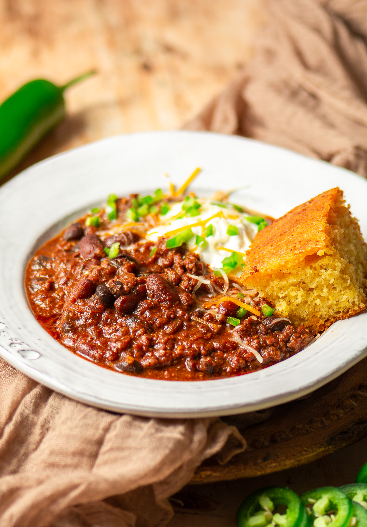 beef and chorizo chili in a bowl topped with sour cream, shredded cheese, diced jalapenos and a slice of cornbread